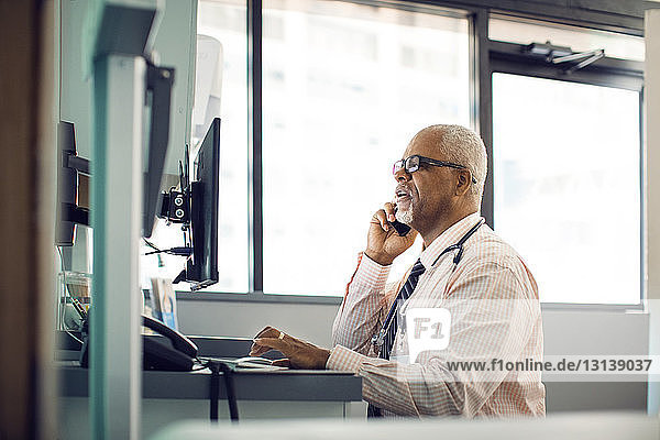 Doctor talking on mobile phone while using desktop computer in hospital
