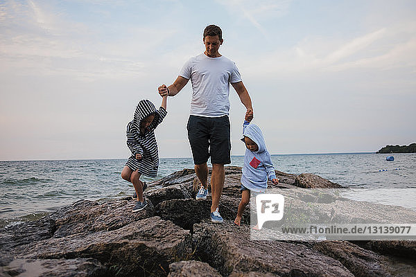 Father and children walking on rocks by sea against sky