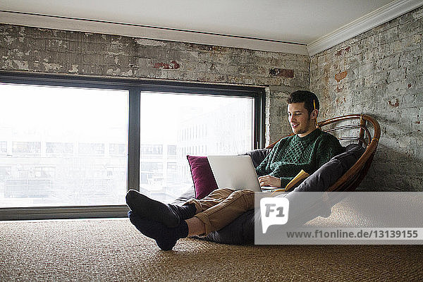 Lächelnder Mann mit Laptop  während er zu Hause auf einem Stuhl am Fenster sitzt
