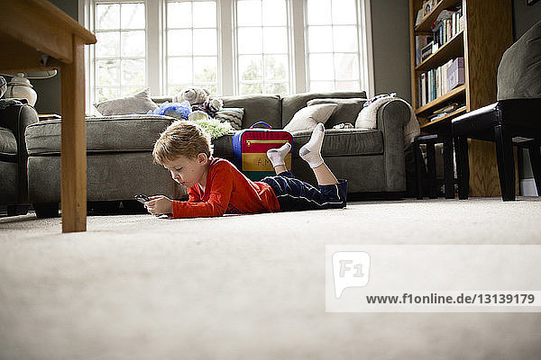 Junge in voller Länge  der ein Smartphone benutzt  während er zu Hause auf dem Teppich liegt