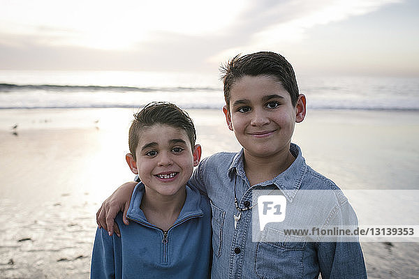 Portrait of smiling brothers standing at beach against sky during sunset