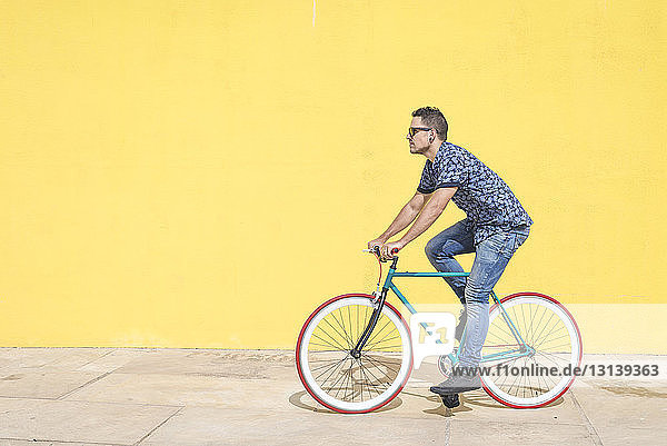Fahrrad fahrender Mann in voller Länge gegen gelbe Wand am Bürgersteig in der Stadt