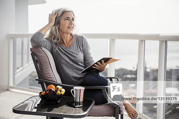 Thoughtful woman holding book while sitting on chair on balcony