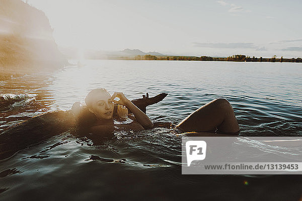 Porträt einer selbstbewussten jungen Frau  die an einem sonnigen Tag im See gegen den Himmel schwimmt