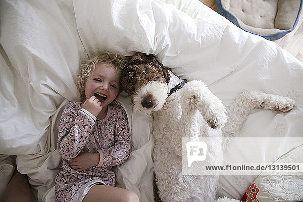 Hochwinkelporträt eines fröhlichen Mädchens  das sich mit seinem Hund zu Hause auf dem Bett entspannt