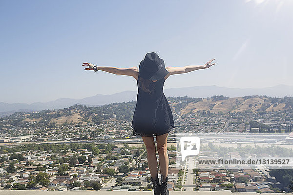 Junge Frau balanciert auf Stützmauer mit Stadt im Hintergrund vor klarem Himmel