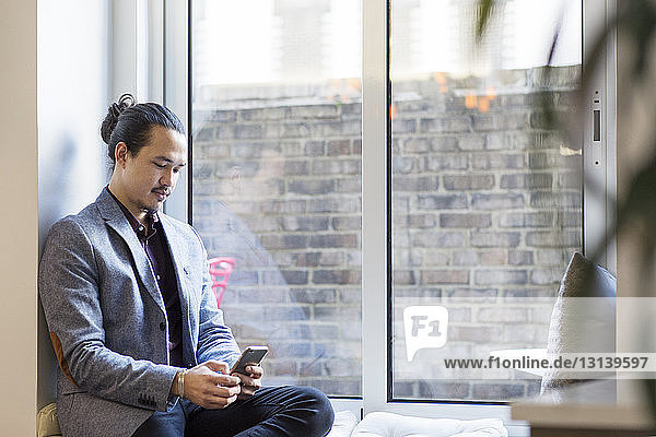 Geschäftsmann benutzt Mobiltelefon  während er im Büro auf einem Fensterplatz in einer Nische sitzt
