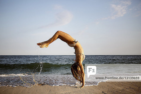 Seitenansicht einer Frau  die am Strand einen Rückwärtssalto gegen den Himmel macht