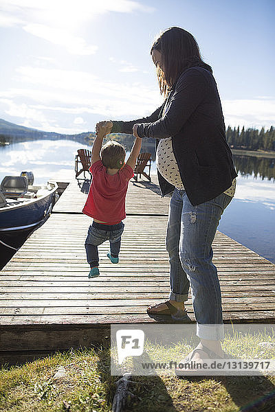 Full length of pregnant mother holding son's hands while swinging him on wooden pier by lake