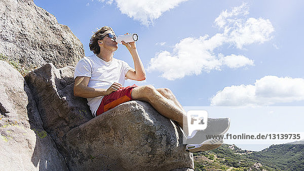 Man drinking water while sitting on rock formation against sky