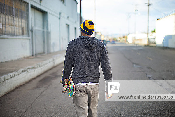 Rückansicht eines Mannes  der ein Skateboard hält und auf der Straße geht