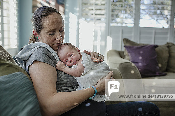 Mutter trägt neugeborenen Sohn  während sie zu Hause auf dem Sofa sitzt