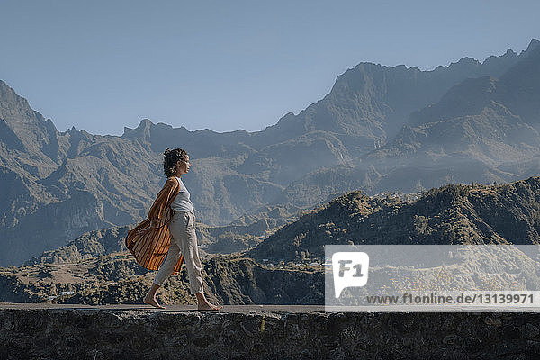 Seitenansicht einer Frau  die auf einer Stützmauer an Bergen vor klarem Himmel entlang läuft