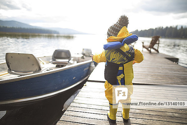 Rear view of baby boy wearing raincoat and life jacket while standing on wooden pier over lake