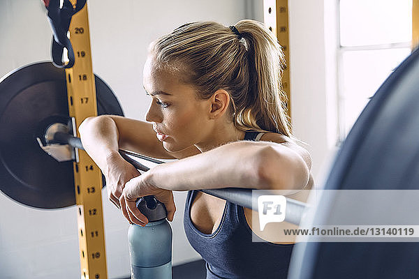 Thoughtful woman leaning on barbell while holding water bottle in gym