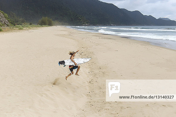 Hochwinkelaufnahme eines hemdlosen Mannes mit Surfbrett  der am Strand springt