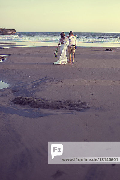 Rückansicht eines frisch vermählten Paares  das bei Sonnenuntergang am Strand bei klarem Himmel spazieren geht