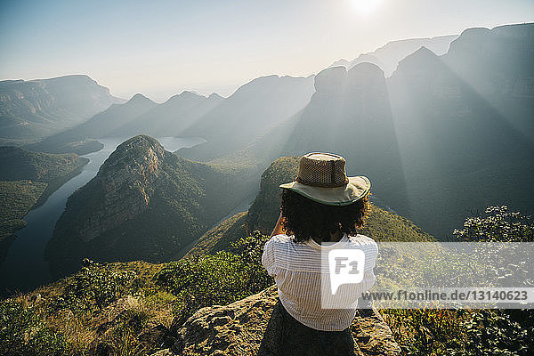 Rear view of woman wearing hat looking at view while sitting on mountain against sky during sunny day