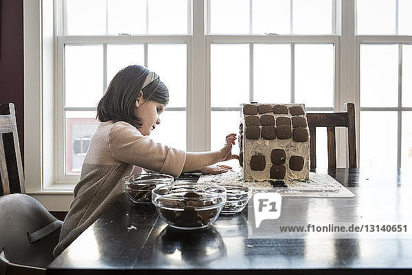 Seitenansicht eines Mädchens  das zu Hause Lebkuchenhaus auf dem Tisch vor dem Fenster macht