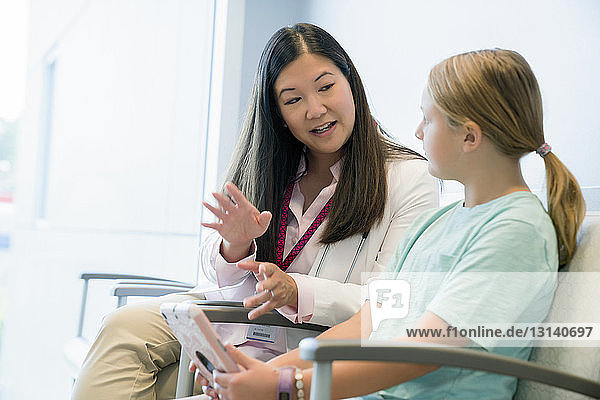 Kinderarzt spricht mit Mädchen  während er im Wartezimmer des Krankenhauses auf einem Stuhl sitzt