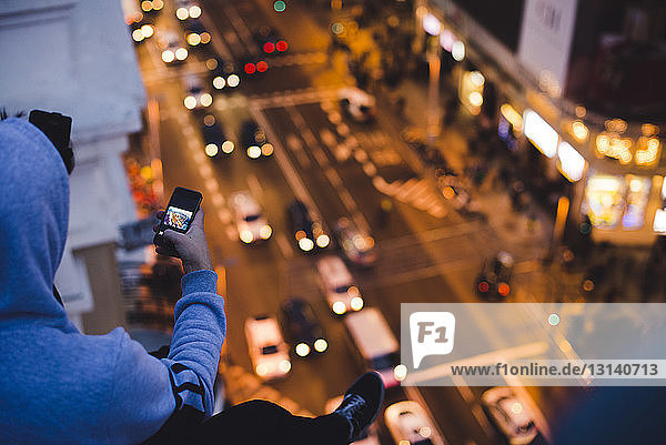 Hochwinkelaufnahme eines Mannes  der eine beleuchtete Straße fotografiert  während er nachts auf der Terrasse eines Gebäudes sitzt