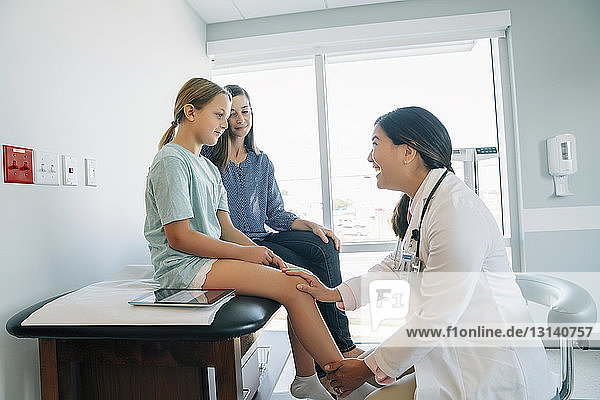 Mother looking at cheerful pediatrician checking daughter's knee joint in medical examination room