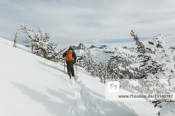 Rückansicht eines Wanderers mit Rucksack und Skistöcken beim Gehen auf einem schneebedeckten Berg