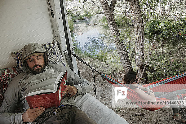 Blick von oben auf den Freund  der im Wohnmobil ein Buch liest  während sich die Freundin in der Hängematte im Wald entspannt