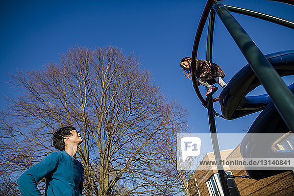 Niedrigwinkelansicht eines Vaters  der seine Tochter beim Klettern auf Außenspielgeräten auf dem Spielplatz beobachtet