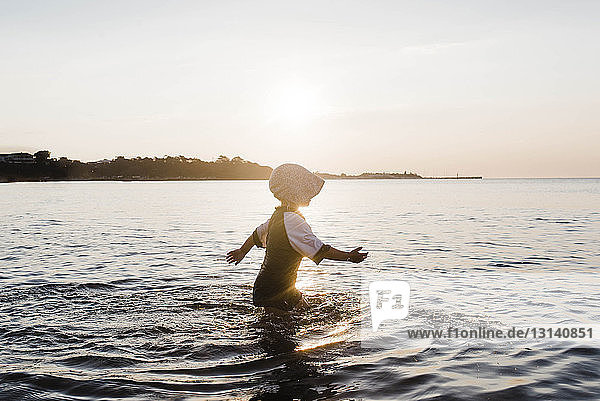 Seitenansicht eines Mädchens mit Mütze beim Spaziergang im Meer gegen den Himmel bei Sonnenuntergang