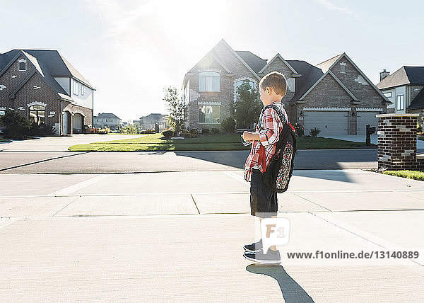 Side view of boy with backpack standing on driveway against sky