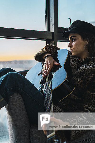 Nachdenkliche junge Frau spielt Gitarre  während sie zu Hause auf dem Sofa vor dem Fenster sitzt