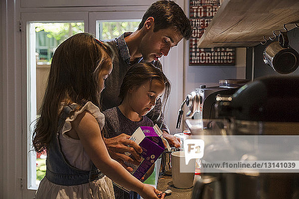 Töchter stehen mit Vater und gießen Milch in Becher an der Küchentheke