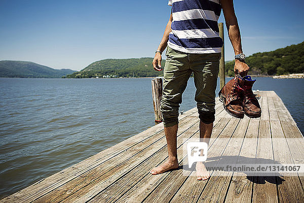 Niedriger Teil eines Mannes mit Schuhen in der Hand  der auf einem Steg über dem See steht