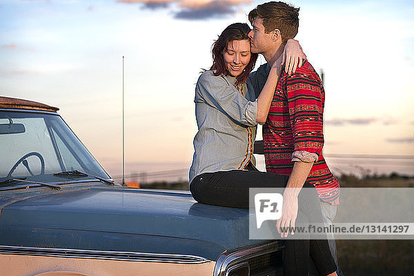 Freundin umarmt ihren Freund  während sie auf der Motorhaube des Fahrzeugs gegen den Himmel sitzt