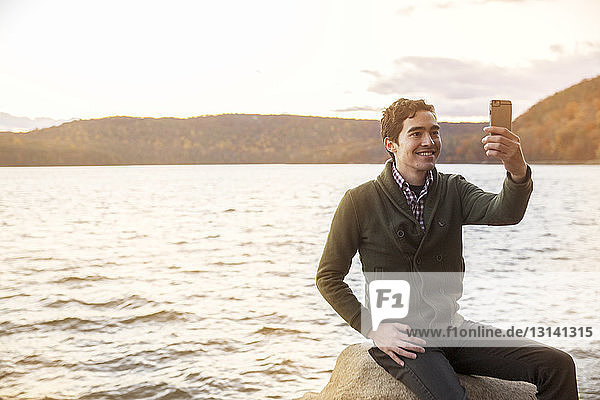 Man taking selfie while sitting on rock against lake