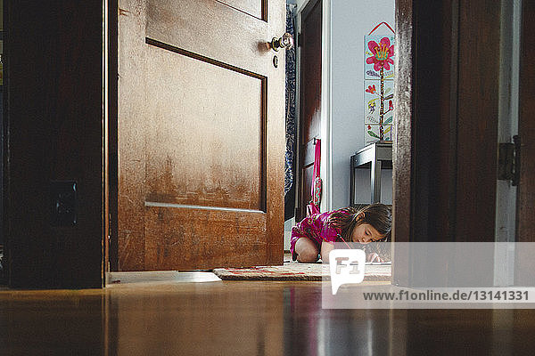 Mädchen macht Hausaufgaben  während sie auf einem Teppich sitzt und durch die Tür zu Hause gesehen wird