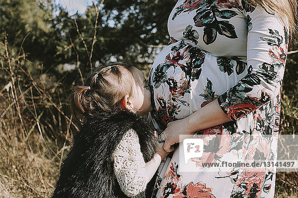 Loving daughter kissing pregnant mother's belly while standing at forest