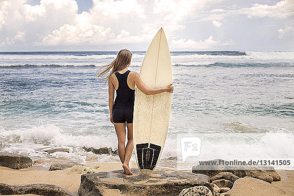 Rückansicht einer Frau  die ein Surfbrett in der Hand hält  während sie an einem felsigen Ufer am Strand steht