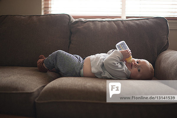 Portrait of baby boy drinking milk from bottle while lying on sofa at home