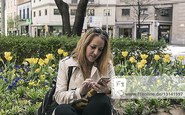 Geschäftsfrau benutzt Mobiltelefon  während sie an Pflanzen in der Stadt sitzt