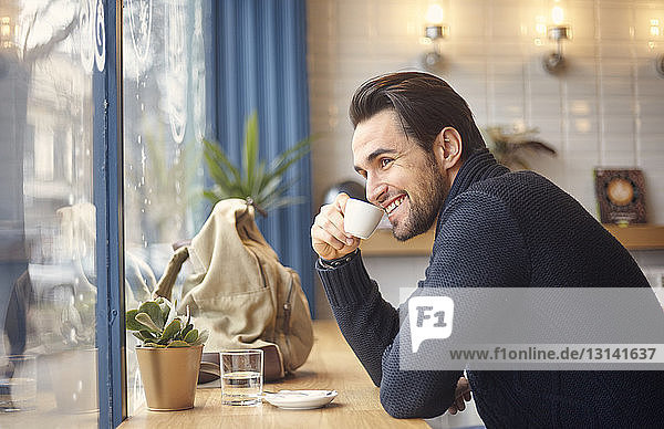 Side view of smiling man looking through window while having coffee by window at cafe