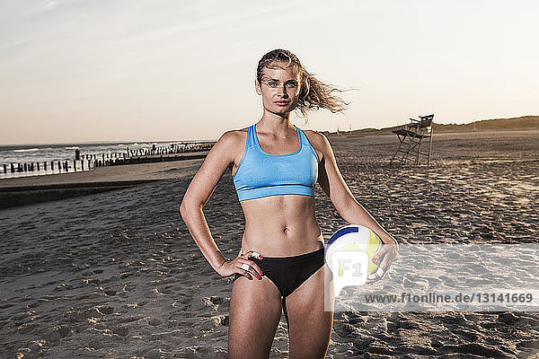 Confident woman holding volleyball and standing at beach