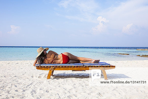 Rückansicht einer Frau  die einen Bikini trägt  während sie am sonnigen Tag auf einer Strandbank gegen den Himmel liegt