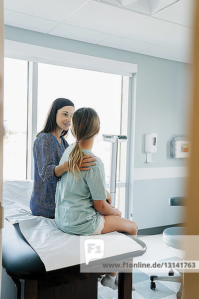 Mother consoling daughter while sitting on examination table seen through doorway