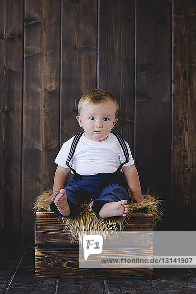 Portrait of cute baby boy sitting on crate against wooden wall