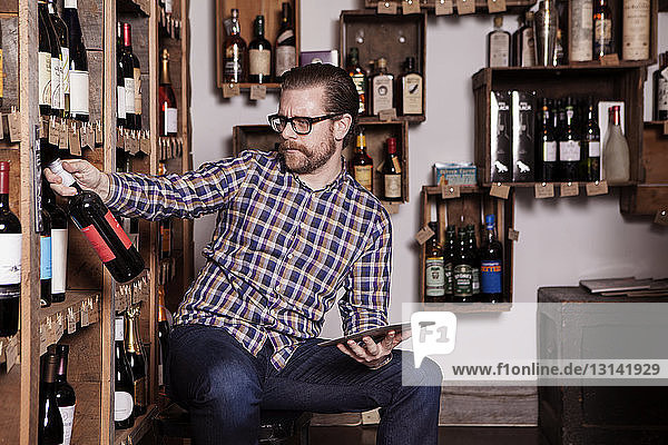 Mature male small business owner reading label on wine bottle while holding tablet computer in shop
