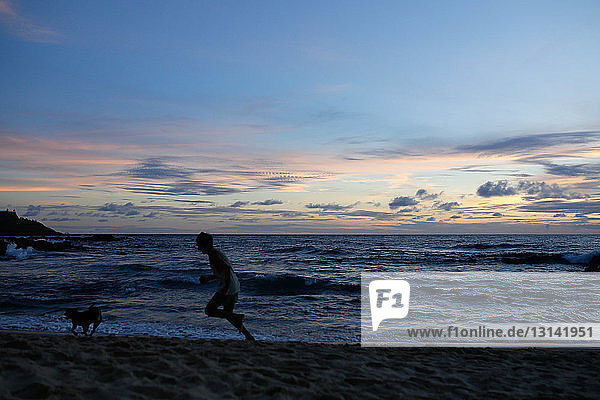 Side view of man and dog running on shore at beach against sky during sunset