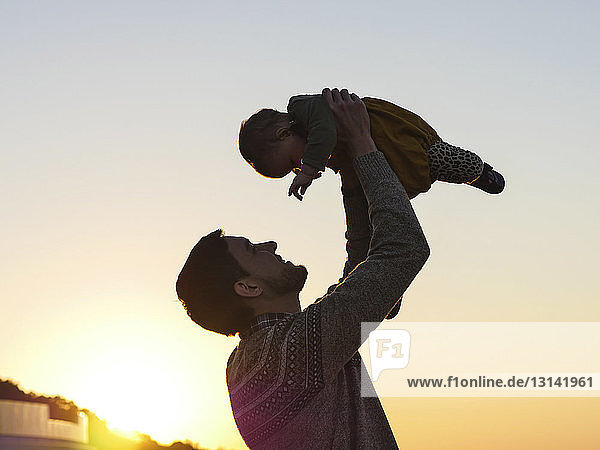 Tiefwinkelansicht eines glücklichen Vaters  der seine Tochter aufhebt  während er bei Sonnenuntergang am klaren Himmel steht