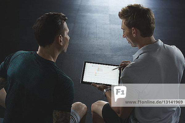Hochwinkelansicht des Trainers  der dem Kunden einen Tablet-PC zeigt  während er im Fitnessstudio sitzt